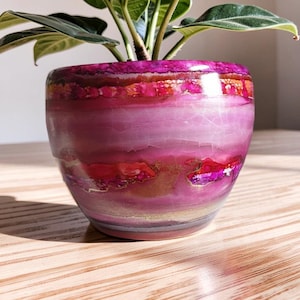 May include: A round ceramic planter featuring a vibrant, multi-layered design in shades of magenta, hot pink, and deep red. The surface displays a glossy, crackled finish with horizontal bands of colour and subtle gold-toned metallic accents throughout.