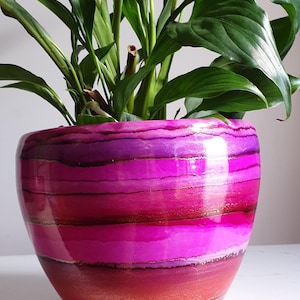 May include: A round ceramic plant pot featuring a glossy finish with horizontal bands of vibrant magenta, deep purple, and reddish-orange colours, accented by thin, shimmering gold lines separating each colour layer, containing a green peace lily plant.