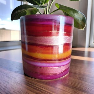 May include: A cylindrical plant pot featuring a glossy, multi-coloured horizontal striped pattern in shades of magenta, deep red, bright orange, and shimmering gold. A green-leafed plant emerges from the top of the smooth, rounded container.