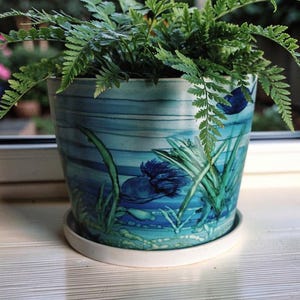 May include: A cylindrical ceramic plant pot featuring a hand-painted underwater scene with shades of blue and teal, depicting aquatic plants and a dark blue fish. The pot sits on a matching white circular saucer and contains a green fern plant.