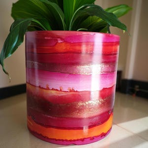May include: A cylindrical plant pot featuring a vibrant, abstract horizontal striped pattern in shades of hot pink, magenta, deep red, and bright orange, accented with shimmering gold metallic bands throughout the glossy, smooth surface.