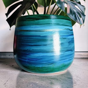 May include: A cylindrical ceramic planter featuring a smooth, glossy finish with horizontal bands of colour transitioning from deep forest green at the top and bottom to vibrant shades of turquoise and royal blue in the centre.