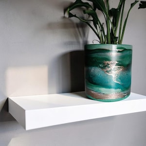 May include: A cylindrical ceramic planter featuring a marbled pattern of deep emerald, teal, and light green hues with metallic gold accents. The planter holds a green leafy plant and sits on a smooth, white rectangular floating wall shelf.
