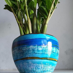 May include: A decorative ceramic vase, hand-painted with blue hues and a textured, wave-like design. The vase contains a green plant with multiple stems and leaves. The vase's design features horizontal bands of varying shades of blue.