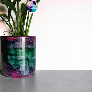 May include: A cylindrical plant pot featuring a vibrant alcohol ink design with swirling patterns of deep green, purple, and magenta. A green leafy plant grows from the top, and a circular logo with the text vrart creation is visible.