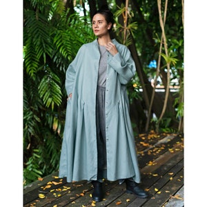 Puede incluir: Abrigo largo azul claro con cuello mandarín y cierre de botones. El abrigo tiene pliegues en la cintura y mangas largas. La persona lleva una camisa y pantalones grises, y botas negras. El fondo presenta follaje verde.