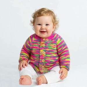 May include: A baby wearing a colorful, striped knit cardigan with wooden buttons. The sweater features vibrant hues of pink, green, and yellow. The baby is also wearing white pants.