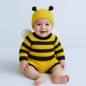 Può includere: Un bambino sorridente indossa un costume da ape a maglia a righe gialle e nere. L'outfit include un cappello con antenne e piccole ali bianche. Il bambino è seduto e guarda direttamente la fotocamera.