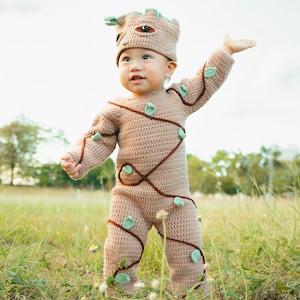 May include: A tan crochet baby costume resembling a tree-like character, complete with a matching hat. The outfit features brown vine-like accents and green leaf details. The baby is standing in a grassy field, arms outstretched.