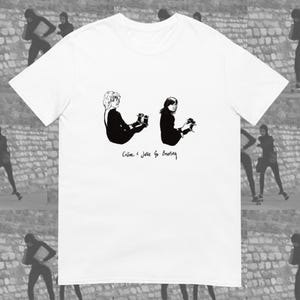 Puede incluir: Camiseta blanca con una ilustración en blanco y negro de dos figuras sentadas, con el texto "Celine + Julie Go Boating". Las figuras visten ropa oscura.