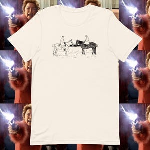 May include: Cream-colored t-shirt featuring a black and white illustration of two people on horseback facing each other. The shirt has a classic crew neck and short sleeves. The background includes a repeating pattern of a person holding a gun.
