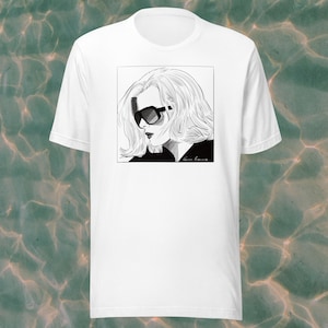 Pode incluir: Camiseta branca com uma ilustração em preto e branco de uma mulher usando óculos de sol. A ilustração é enquadrada em um quadrado e a assinatura do artista é visível no canto inferior direito.