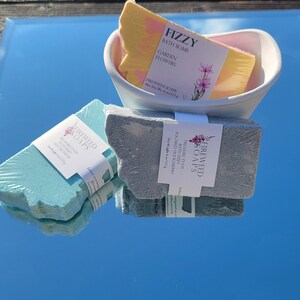 May include: Three bath products from Fireweed Soaps. A yellow bath bomb with the text "Fizzy Bath Bomb Garden Flowers" in a white bowl. A blue bath fizzy with the text "Treasure State Sugared Huckleberry Bath Fizzy" and a blue bath bar with the text "Treasure State Bath Fizzy Sugared Huckleberry".