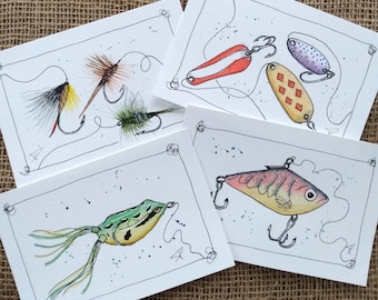 May include: Four watercolor fishing lure illustrations on white cards. The lures include a frog, a fish, and various hooks with colorful feathers. Each card has a black border and a decorative corner design. The background is a textured, tan fabric.