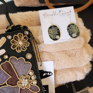 May include: A black and gold embroidered clutch purse with floral designs. The purse is next to a white faux fur fabric and a card with the text "Bramble Grove Boutique" and a pair of gold earrings with pressed flowers.