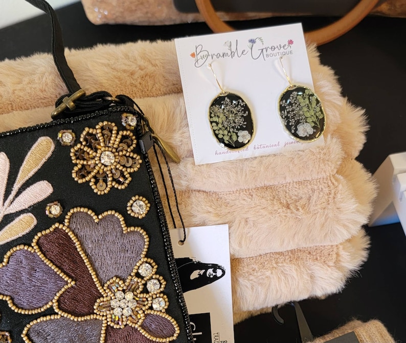 May include: A black and gold embroidered clutch purse with floral designs. The purse is next to a white faux fur fabric and a card with the text "Bramble Grove Boutique" and a pair of gold earrings with pressed flowers.