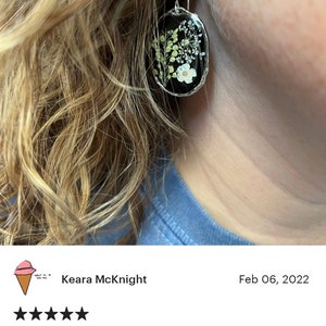 May include: A pair of silver earrings with a black oval-shaped resin setting. The resin contains pressed white and green flowers. The earrings are hanging from a person's ear.
