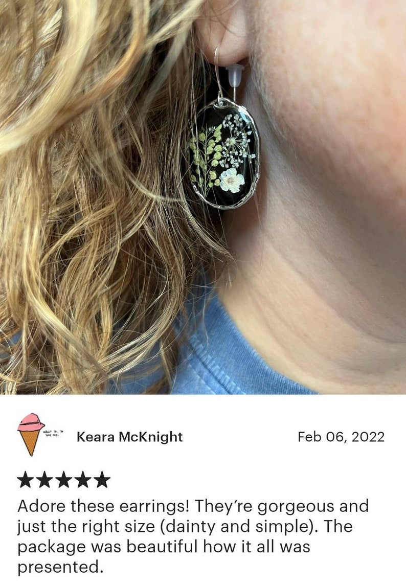 May include: A pair of silver earrings with a black oval-shaped resin setting. The resin contains pressed white and green flowers. The earrings are hanging from a person's ear.