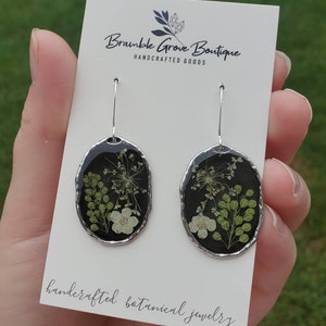 May include: A pair of silver earrings with black resin and pressed white and green flowers. The earrings are oval shaped and have a silver hook. The card reads "Bramble Grove Boutique Handcrafted Goods handcrafted botanical jewelry".