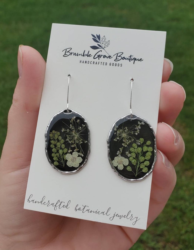 May include: A pair of silver earrings with black resin and pressed white and green flowers. The earrings are oval shaped and have a silver hook. The card reads "Bramble Grove Boutique Handcrafted Goods handcrafted botanical jewelry".