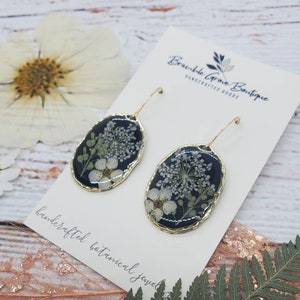 May include: A pair of black and gold oval earrings with dried white flowers and greenery encased in resin. The earrings are handcrafted and have a gold hook closure. The earrings are displayed on a card that reads "Bramble Grove Boutique Handcrafted Goods" and "Handcrafted Botanical Jewelry".
