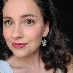 May include: A woman with dark brown hair wearing a white tank top and gold earrings with a pressed flower design.