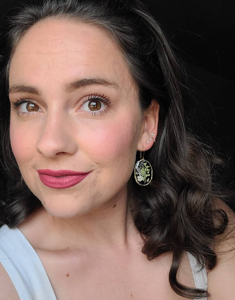 May include: A woman with dark brown hair wearing a white tank top and gold earrings with a pressed flower design.