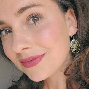 May include: A close-up of a person's face with a pink lip color. The person is wearing gold earrings with a black oval-shaped design featuring dried flowers.