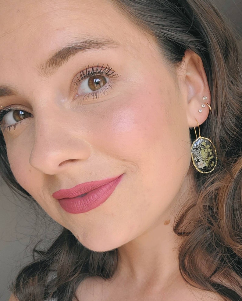 May include: A close-up of a person's face with a pink lip color. The person is wearing gold earrings with a black oval-shaped design featuring dried flowers.