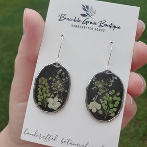 May include: A pair of silver dangle earrings with black resin oval pendants. Each pendant features pressed white and green flowers. The card reads "Bramble Grove Boutique Handcrafted Goods" and "Handcrafted Botanical Jewelry".
