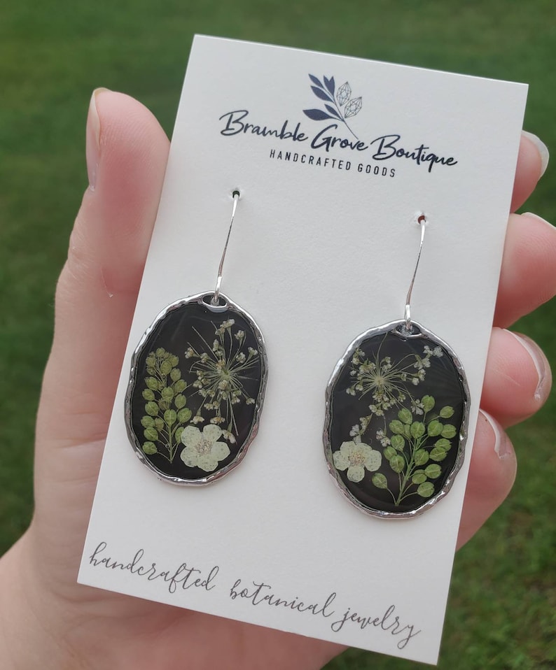 May include: A pair of silver dangle earrings with black resin oval pendants. Each pendant features pressed white and green flowers. The card reads "Bramble Grove Boutique Handcrafted Goods" and "Handcrafted Botanical Jewelry".