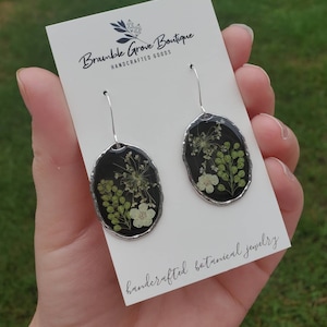 May include: A pair of silver earrings with black oval-shaped resin that encases pressed flowers and greenery. The earrings are handcrafted botanical jewelry. The text "Bramble Grove Boutique Handcrafted Goods" is on the card behind the earrings.