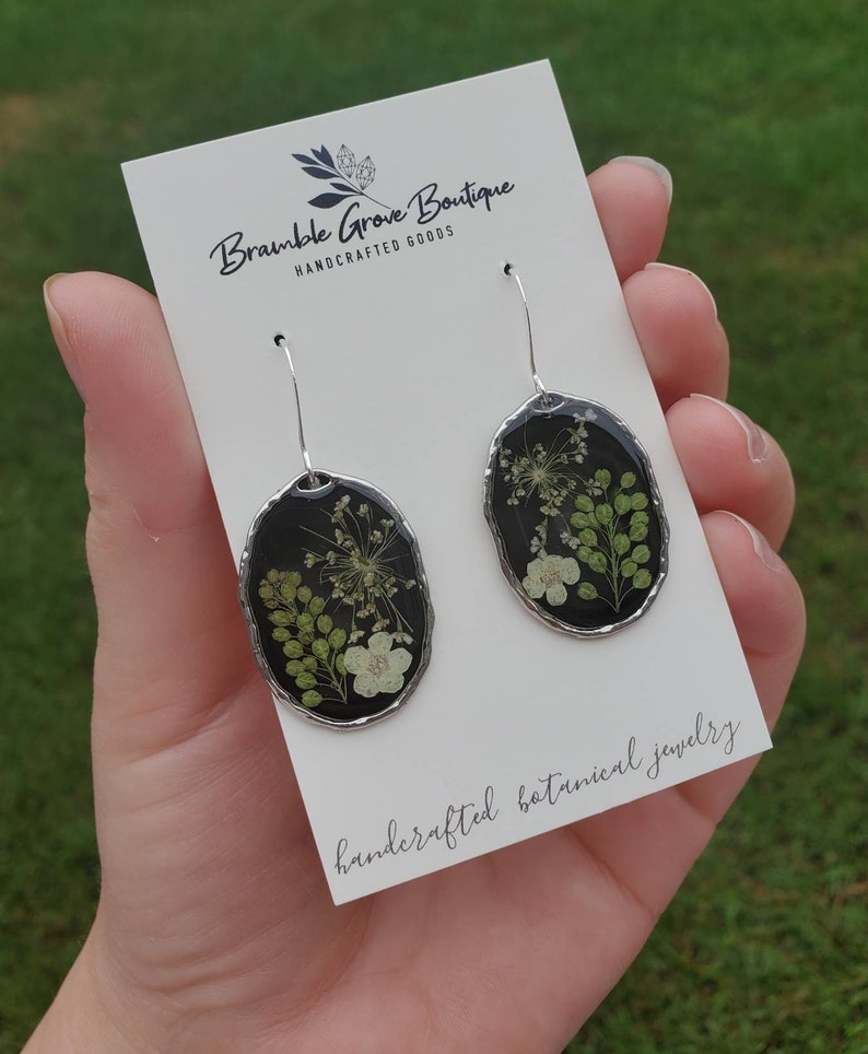 May include: A pair of silver earrings with black oval-shaped resin that encases pressed flowers and greenery. The earrings are handcrafted botanical jewelry. The text "Bramble Grove Boutique Handcrafted Goods" is on the card behind the earrings.
