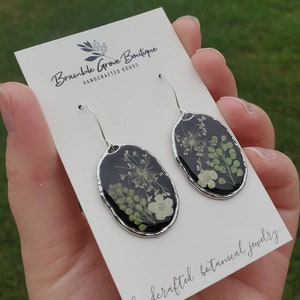 May include: A pair of silver dangle earrings with oval-shaped resin pendants featuring pressed dried flowers in shades of green and white against a black background. The earrings are displayed on a white card with the text "Bramble Grove Boutique Handcrafted Goods" and "handcrafted botanical jewelry".