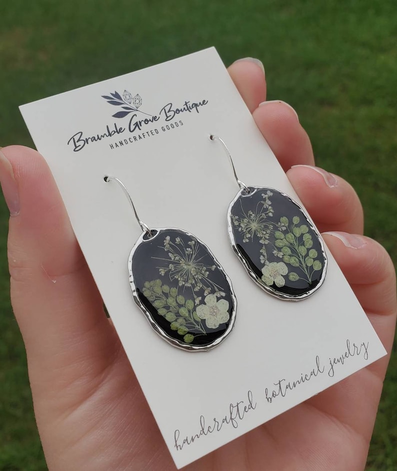 May include: A pair of silver dangle earrings with oval-shaped resin pendants featuring pressed dried flowers in shades of green and white against a black background. The earrings are displayed on a white card with the text "Bramble Grove Boutique Handcrafted Goods" and "handcrafted botanical jewelry".