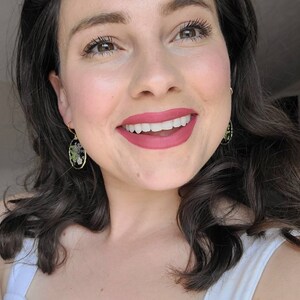 May include: A woman with long dark brown hair is smiling and wearing a white tank top. She is wearing earrings with a black oval background and a floral design.