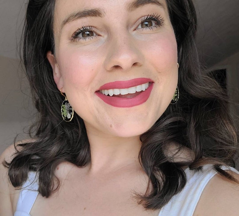 May include: A woman with long dark brown hair is smiling and wearing a white tank top. She is wearing earrings with a black oval background and a floral design.
