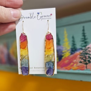 Könnte beinhalten: Regenbogenfarbene gepresste Blumenohrringe mit goldenen Haken. Die Ohrringe sind rechteckig und zeigen eine Vielzahl von Farben, darunter Rot, Orange, Gelb, Grün, Blau und Lila.