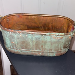 May include: An oval copper basin featuring a weathered exterior with a prominent green patina finish. The interior displays a polished, warm metallic copper tone. The vessel includes small metal handles on each side and a horizontal ridge.