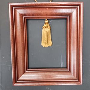 May include: A rectangular, empty picture frame with a dark wood finish. A gold tassel hangs from the top center. The frame has a layered design, and the background is a dark gray.