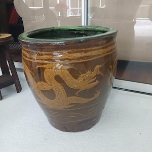 May include: A large, brown ceramic planter with a dragon design. The planter has a wide, cylindrical shape with a green rim and a textured surface. The dragon is a light brown color and is carved into the side of the planter.