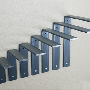 May include: A set of seven dark gray metal shelf brackets arranged in a descending staircase pattern. Each bracket is L-shaped with a flat top and a vertical support, secured to the wall with screws. The brackets are designed for supporting shelves.