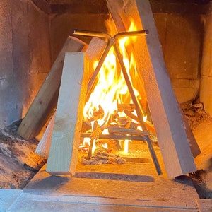 May include: A metal fire grate with a triangular shape is placed inside a brick fireplace. The grate is holding burning logs and kindling, creating a warm orange and yellow flame.