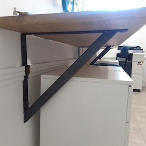 Op de afbeelding: Een zwarte metalen plankbeugel is bevestigd aan een witte muur en ondersteunt een houten plank. De plank is gemonteerd boven een wit bureau met laden.