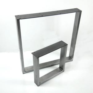 Pode incluir: Duas pernas de mesa de metal, uma maior que a outra, são mostradas em um estúdio branco. As pernas são feitas de tubos de metal quadrados e têm uma aparência crua e inacabada.