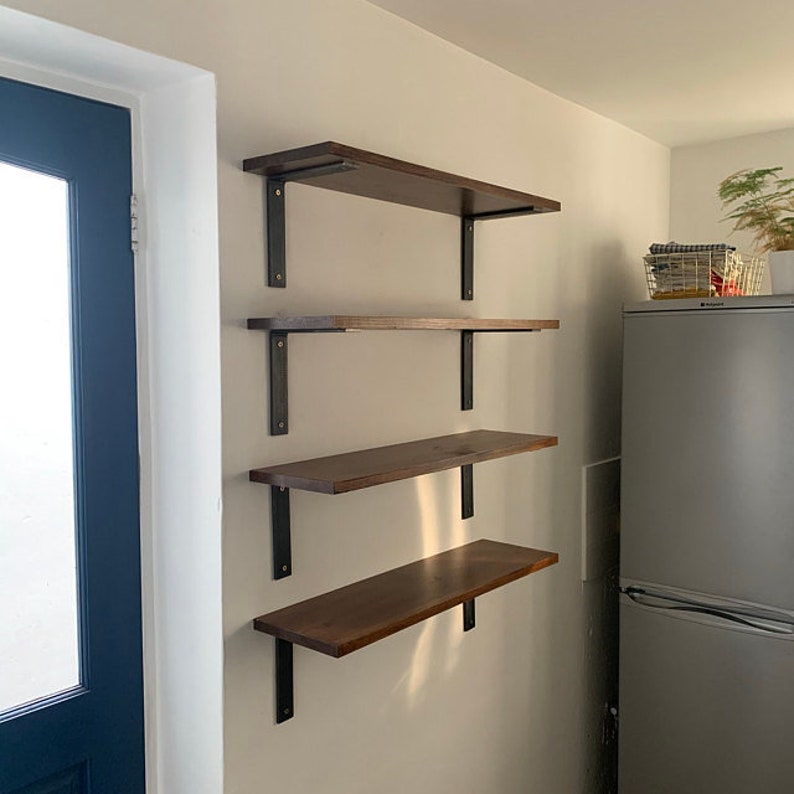 May include: Four wooden shelves mounted on a wall with black metal brackets. The shelves are a dark brown color and are different lengths. The shelves are mounted on a light beige wall. The shelves are in a kitchen or pantry.