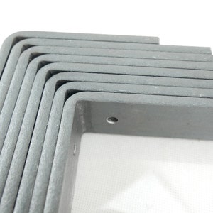 May include: A stack of gray, L-shaped corner brackets. Each bracket has a rounded corner and a pre-drilled hole. The brackets are made of a textured material and are stacked on top of each other.