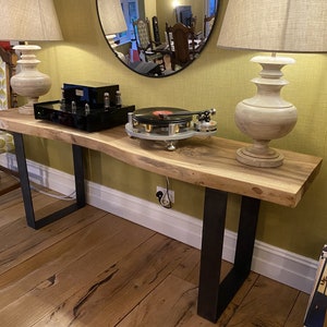 May include: A long, live edge wood table with black metal legs. The table has a natural wood finish and is topped with a record player, a black amplifier, and two lamps with white shades.