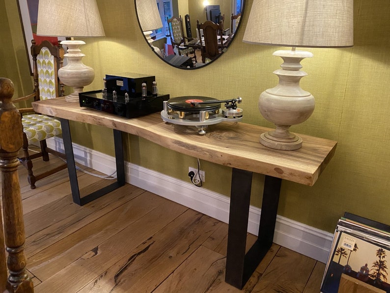 May include: A long, live edge wood table with black metal legs. The table has a natural wood finish and is topped with a record player, a black amplifier, and two lamps with white shades.