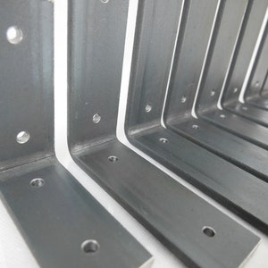 May include: Several L-shaped metal brackets with pre-drilled holes are arranged in a row. The brackets are made of a dark gray metal and have a matte finish. The design is simple and industrial, suitable for shelving or other support applications.